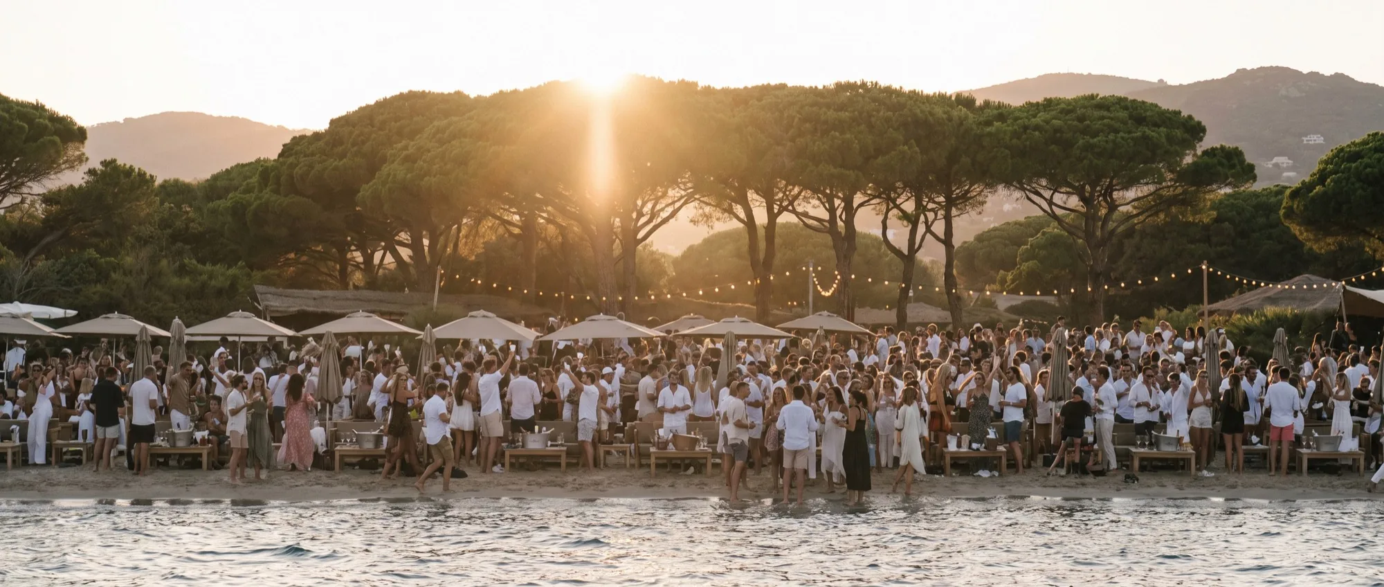 A Riviera beach club in full swing at golden hour, guests dancing on the sand with champagne under string lights.