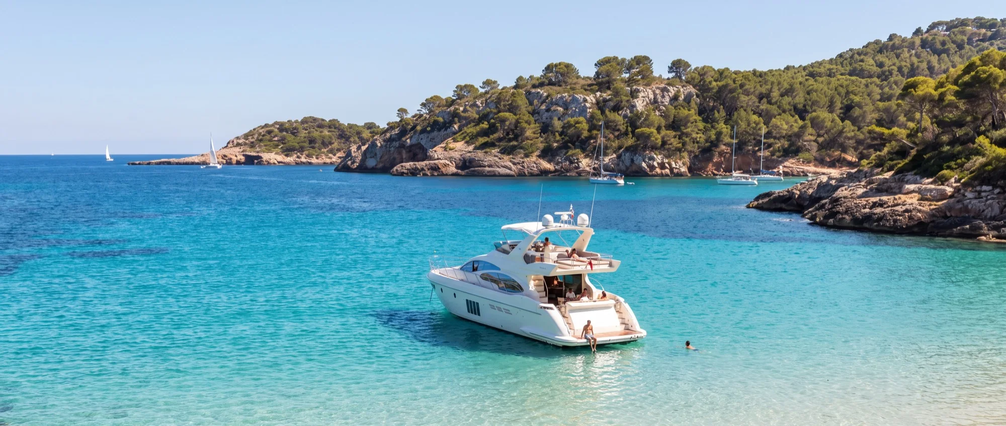 A motor yacht at anchor in a wide turquoise cove, guests aboard and one swimming off the stern.
