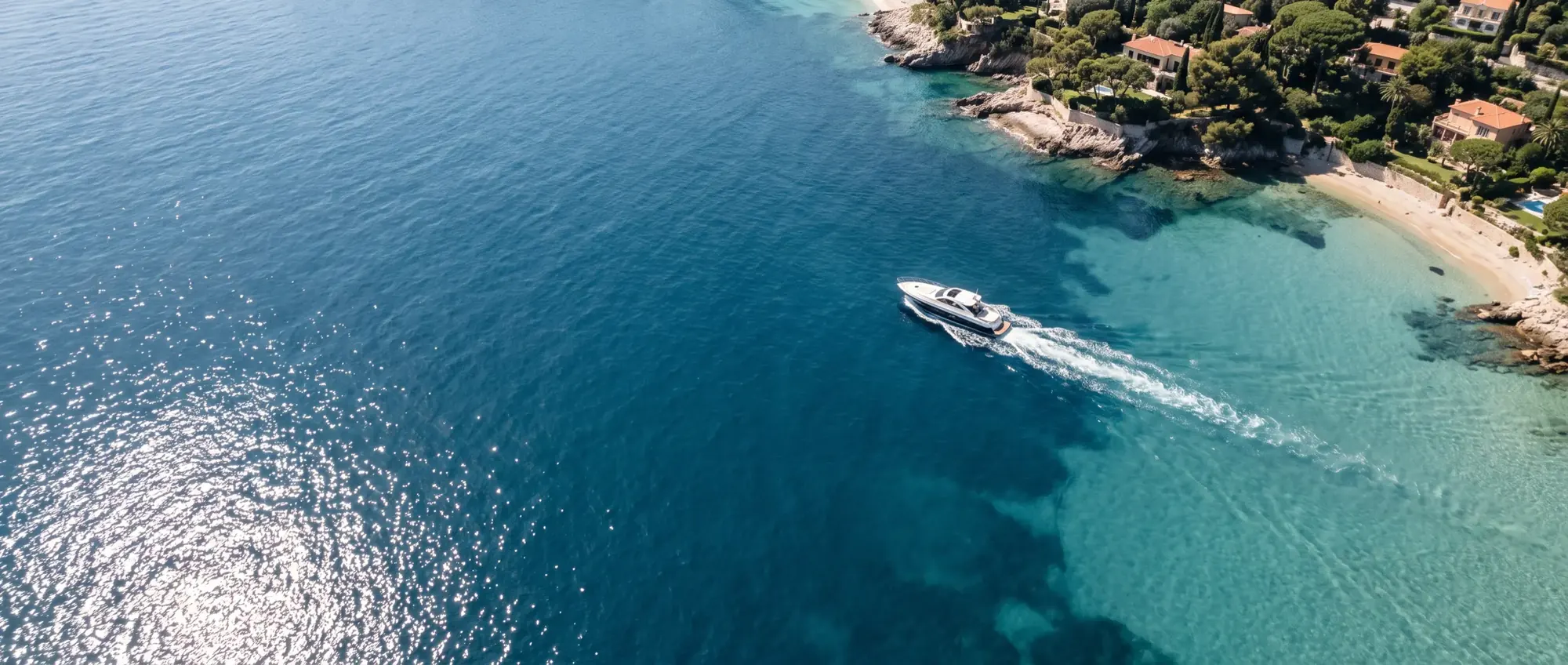 A day yacht crossing sapphire water off the Riviera coast, seen from above, villas and a sandy cove beyond.